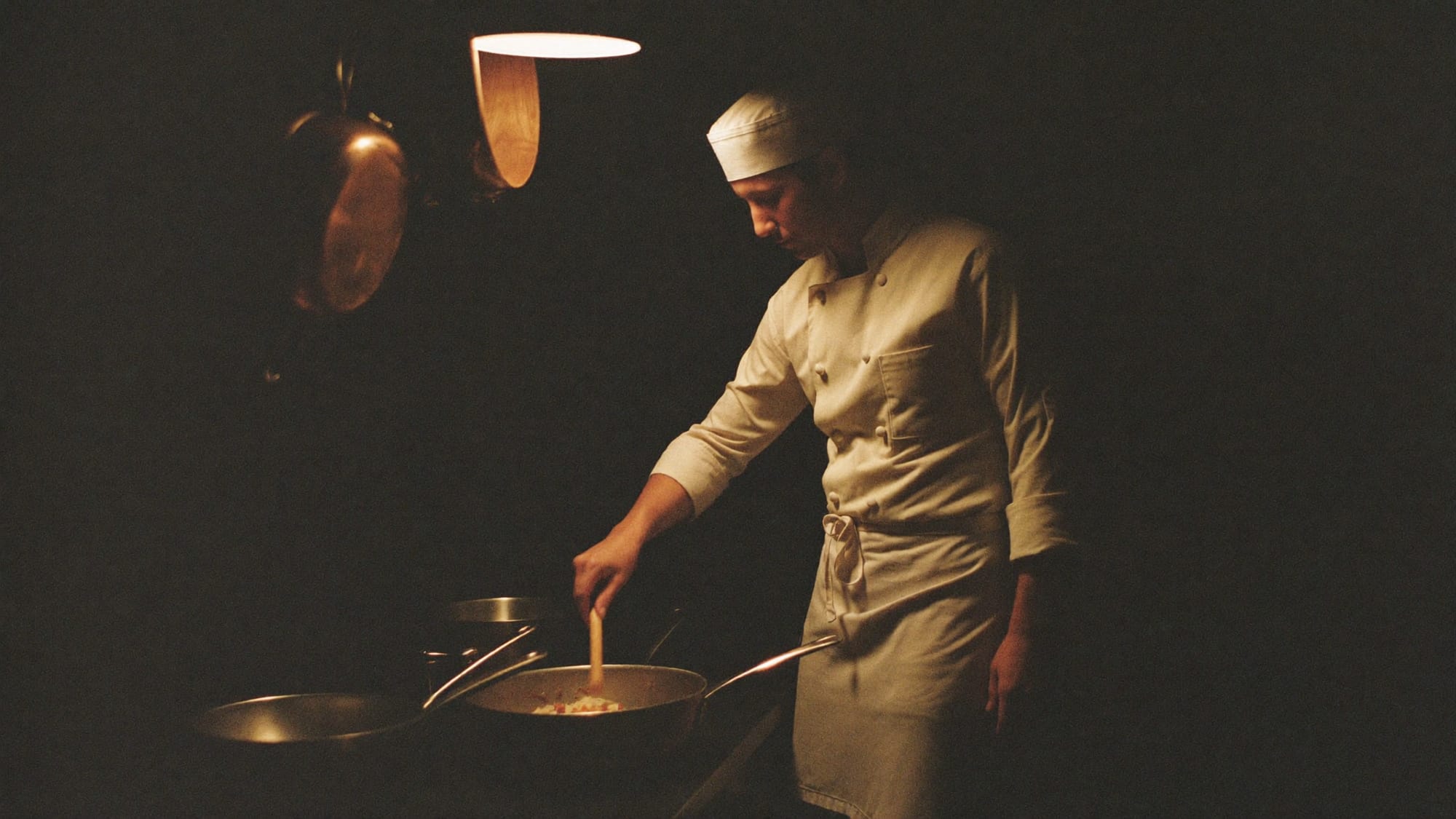 A private chef under a single pendant light in a dark kitchen, copper pans