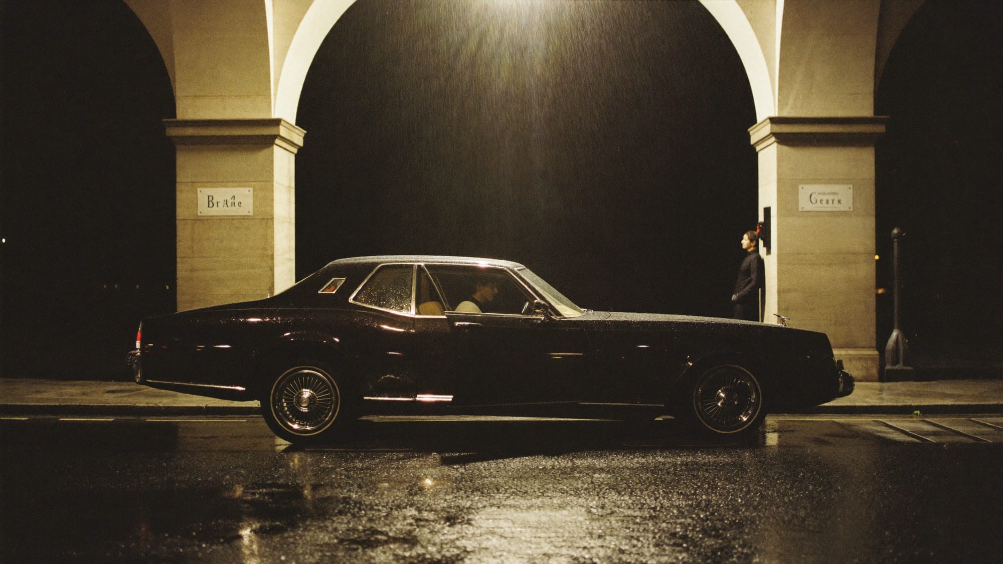 A dark saloon car waiting under a porte-cochère at night, rain on stone