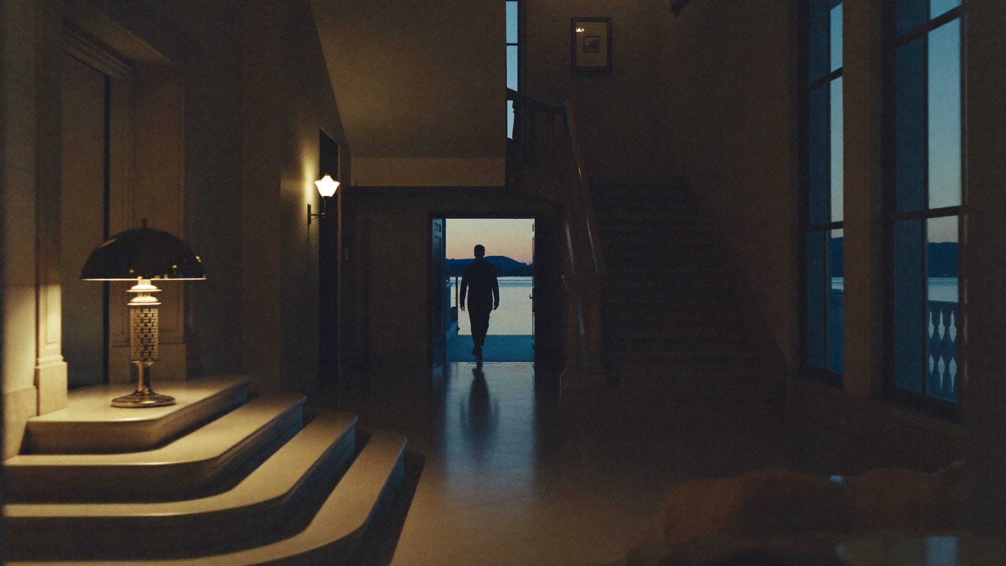The entrance hall of a lakeside villa at dusk, one lamp lit, a figure walking out toward the water