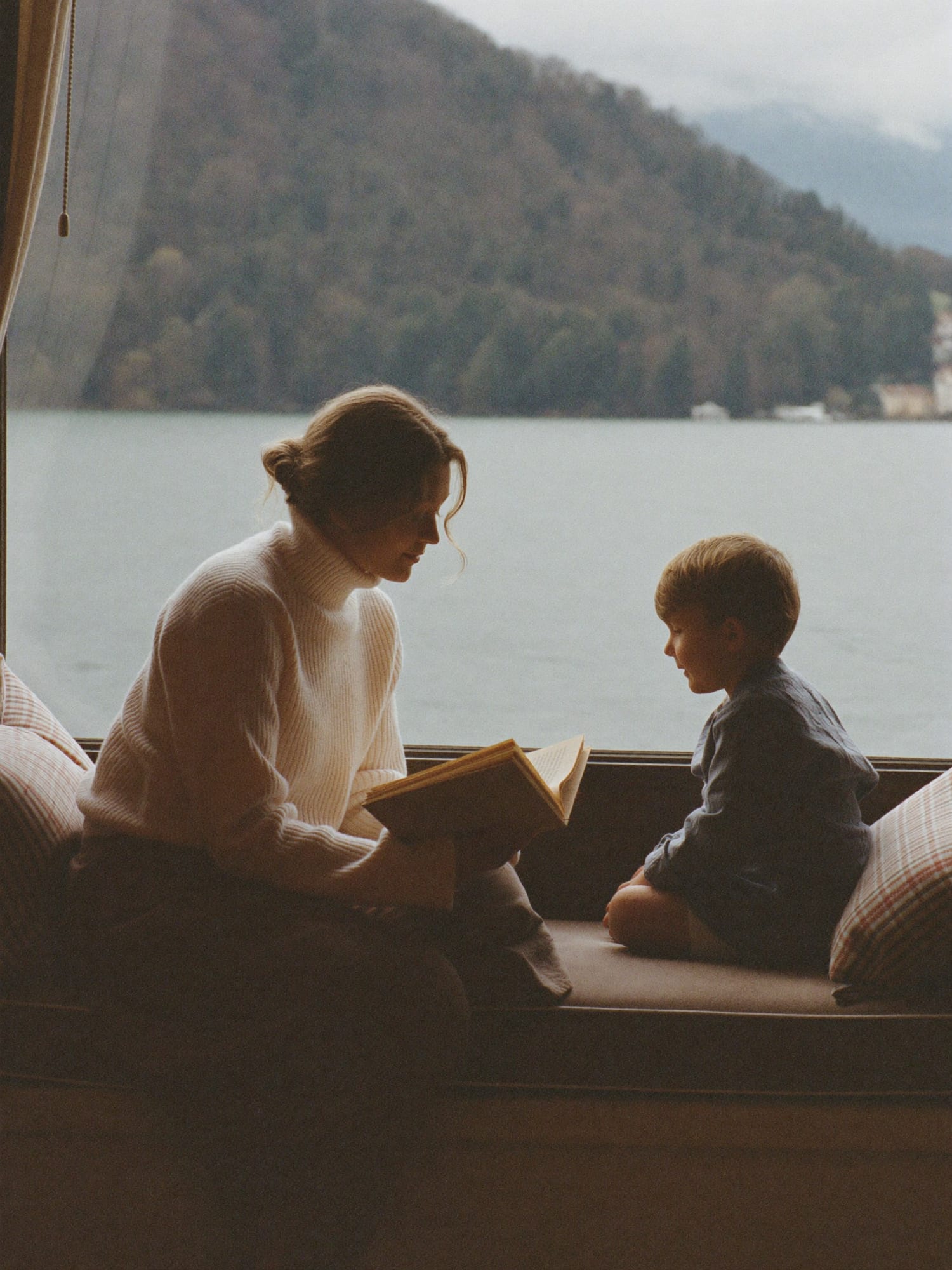 A nanny reading a picture book with a child on a window seat